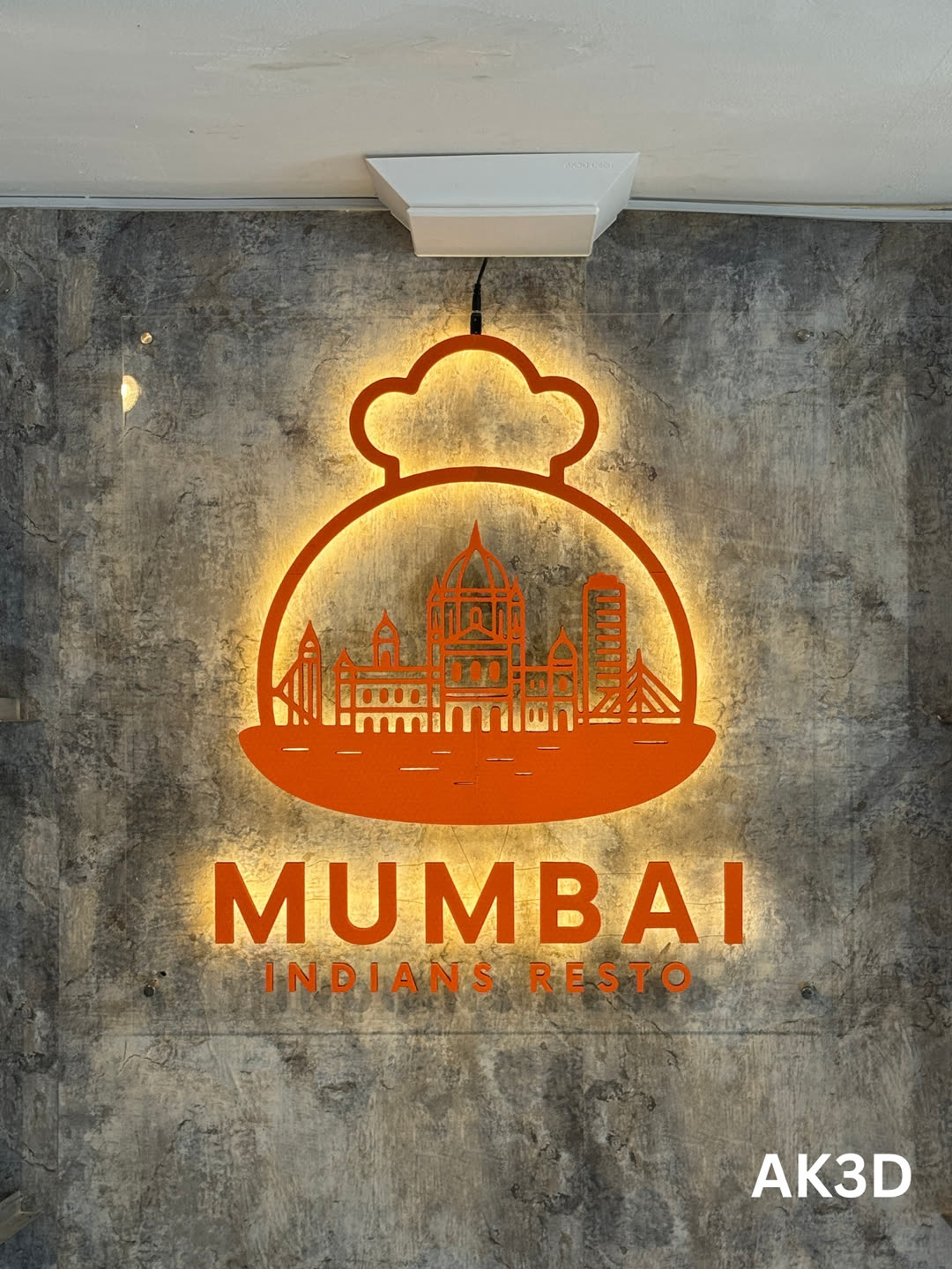 Orange Mumbai Indians Resto logo sign with a skyline under a dome, glowing with warm LED backlight on a grey concrete-look wall
