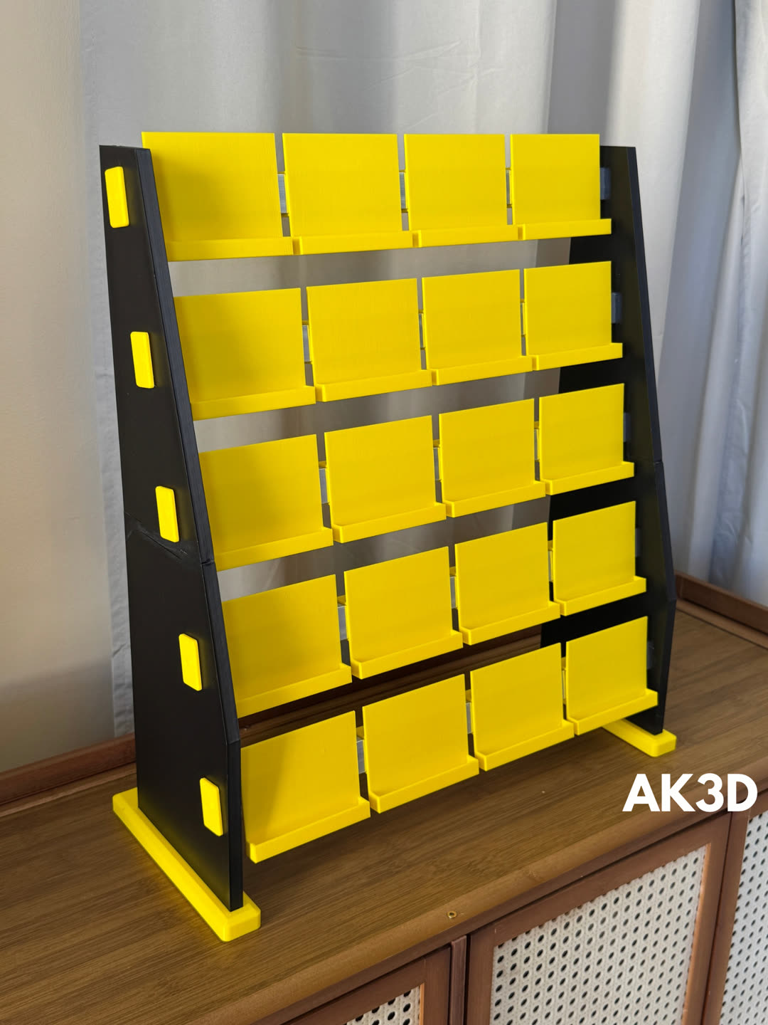 Black 3D printed display stand with twenty yellow sticker trays in five rows, standing on a wooden cabinet